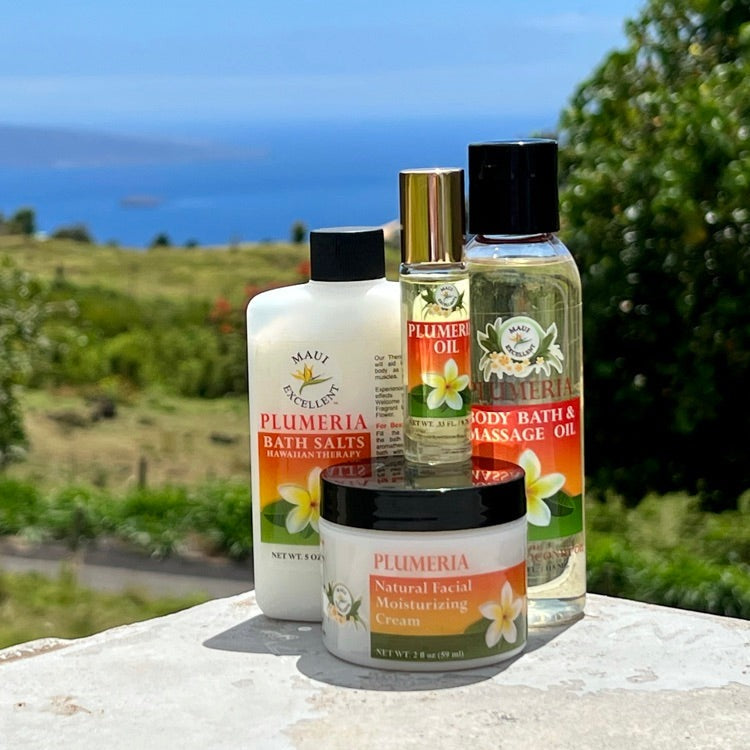 Maui Excellent Plumeria Product Collection on table: Full-sized Essential Oil Bath Salts, Roll-On Oil, Body Bath and Massage Oil, and Natural Facial Moisturizing Cream. Trees, ocean, and distant island in background.