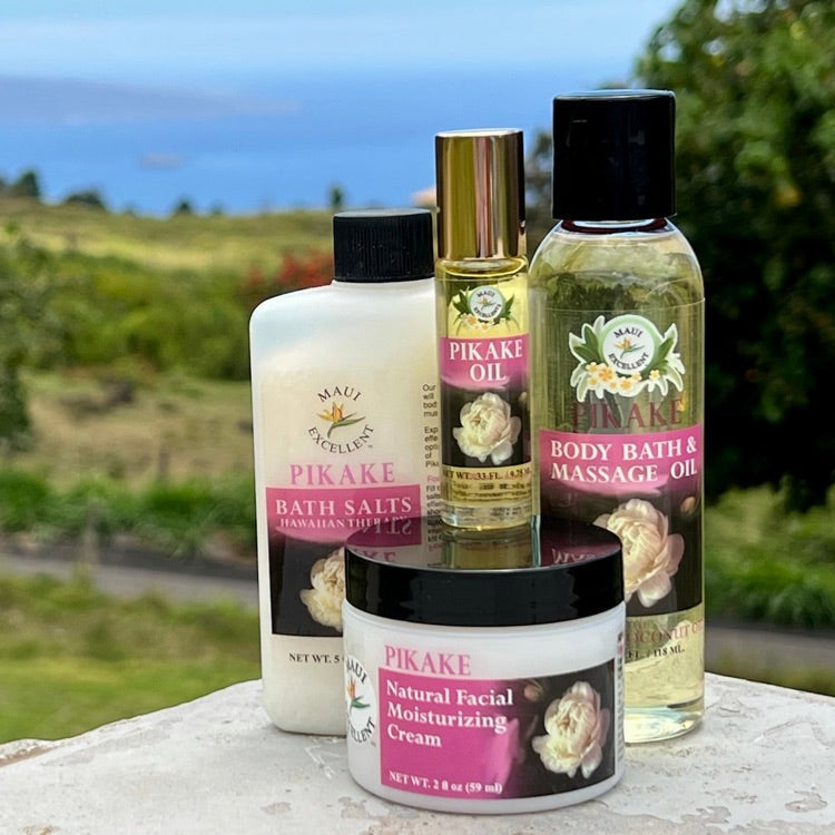 Maui Excellent Pikake Product Collection on table: Full-sized Essential Oil Bath Salts, Roll-On Oil, Body Bath and Massage Oil, and Natural Facial Moisturizing Cream. Trees, ocean, and distant island in background.