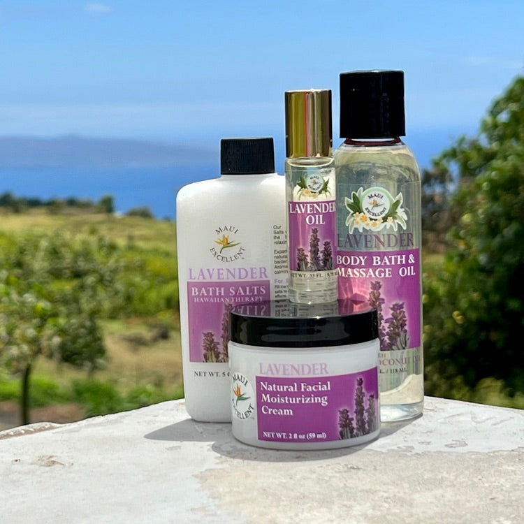 Maui Excellent Lavender Product Collection on table: Full-sized Essential Oil Bath Salts, Roll-On Oil, Body Bath and Massage Oil, and Natural Facial Moisturizing Cream. Trees, ocean, and distant island in background.