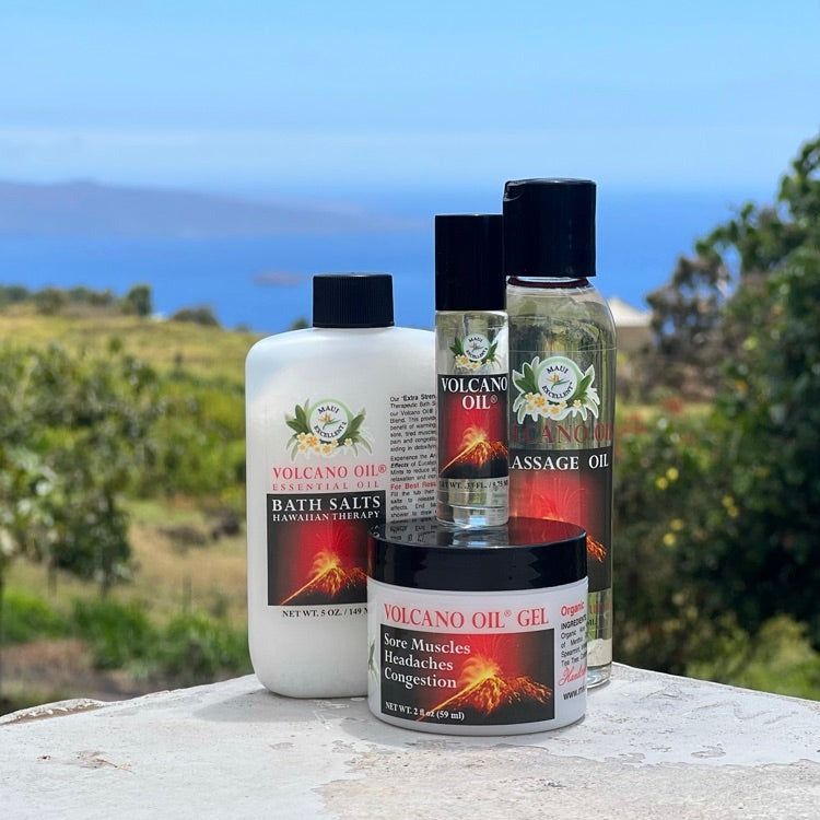 Maui Excellent Volcano Oil Product Collection on table: Essential Oil Bath Salts, Roll-On Oil, Body Bath and Massage Oil, and Volcano Oil Gel. Trees, ocean, and distant island in background.