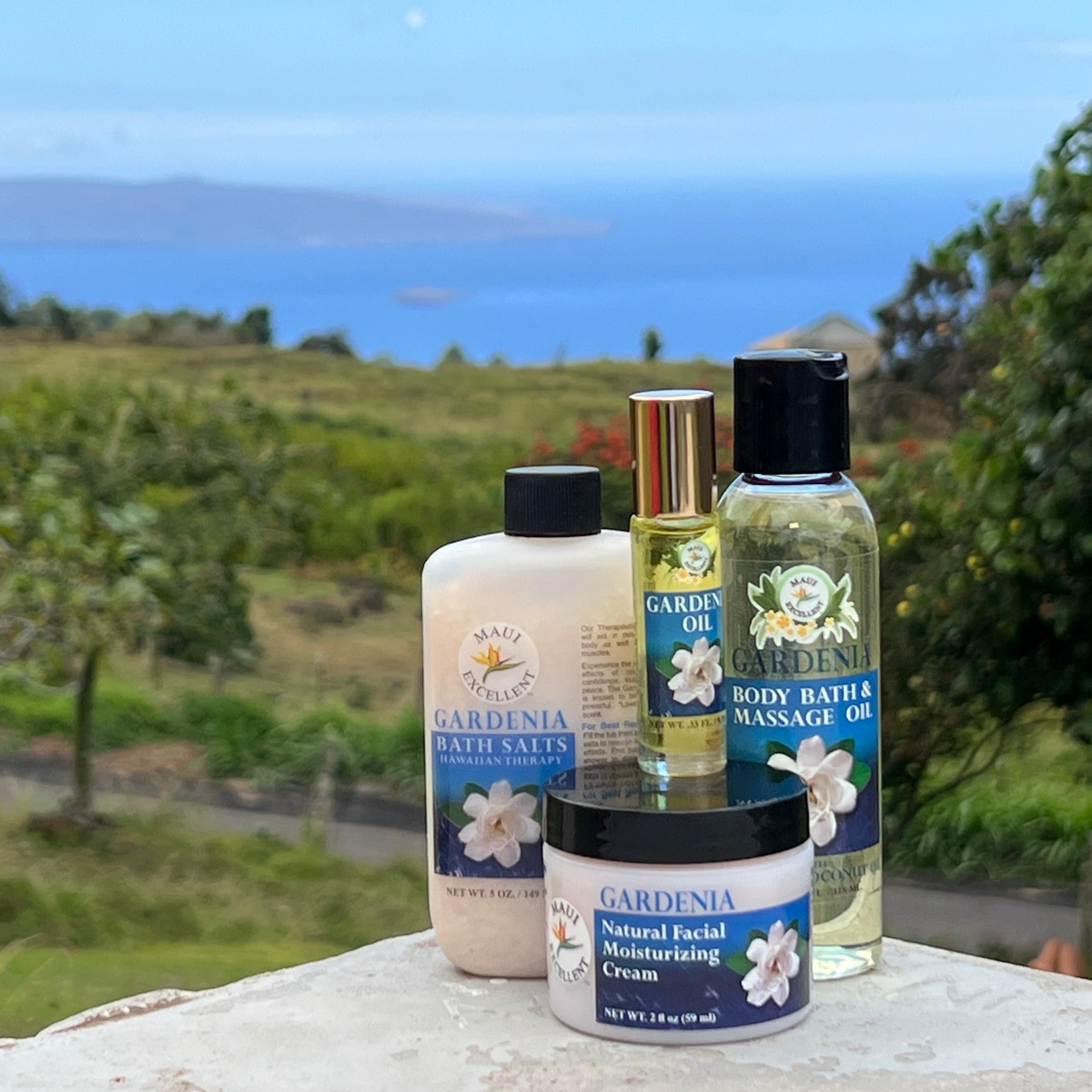 Maui Excellent Gardenia Product Collection on table: Full-sized Essential Oil Bath Salts, Roll-On Oil, Body Bath and Massage Oil, and Natural Facial Moisturizing Cream. Trees, ocean, and distant island in background.
