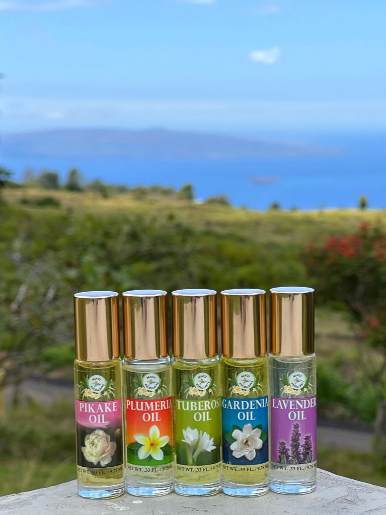 All five Maui Excellent Hawaiian Floral Essential Oil Roll-On bottles lined up in foreground. Green hill, ocean, and island in background.