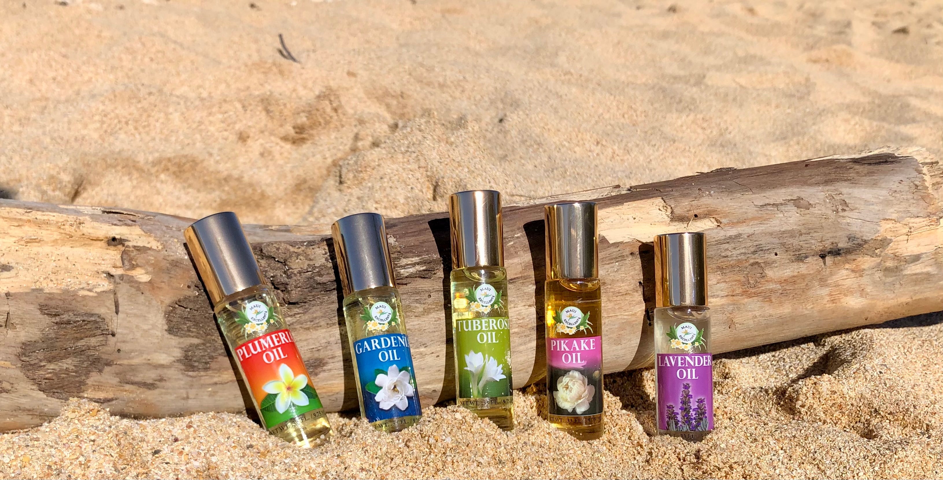 All five Maui Excellent Hawaiian Floral Essential Oil Roll-On bottles are leaning on a small log on the sand. 