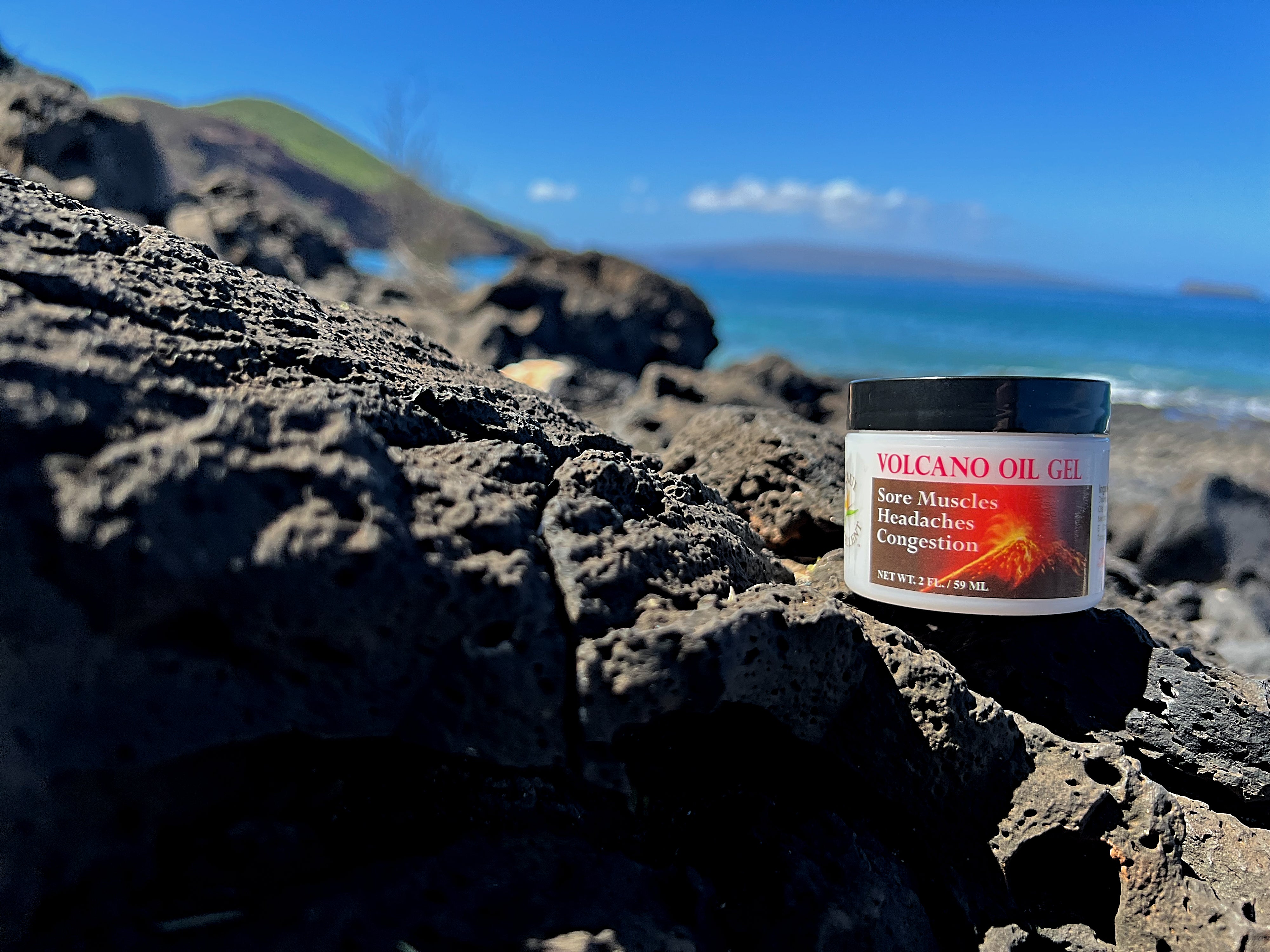 Maui Excellent Volcano Oil Gel atop gray beach rocks. Blue sky, ocean, the Hawaiian Island Kahoolawe, and the Molokini Crater in the distant background.